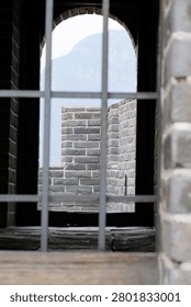 Interior view through iron window bars and a dark arched opening inside a Great Wall of China watchtower, looking out onto grey crenellated parapets and a mountain silhouette against a bright sky, framing an intimate glimpse of the ancient structure
