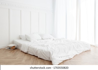 Interior view of white unmade messy bed on a wooden floor in the morning with transparent curtain and sunlight