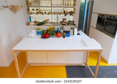 Interior White Kitchen, No People, The Kitchen Has A Table With Food For Cooking, Oven, Built-in Microwave, Yellow Floor, White Walls, Shelves With Accessories