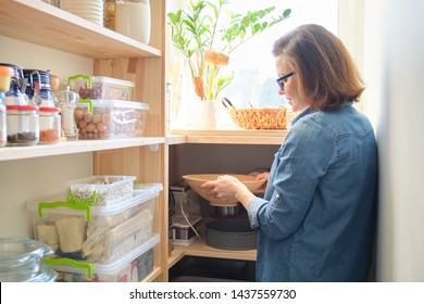 Interior of wooden pantry with products for cooking. Adult woman taking kitchenware and food from the shelves.