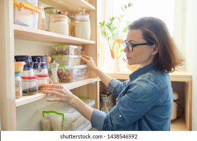 Interior of wooden pantry with products for cooking. Adult woman taking kitchenware and food from the shelves.