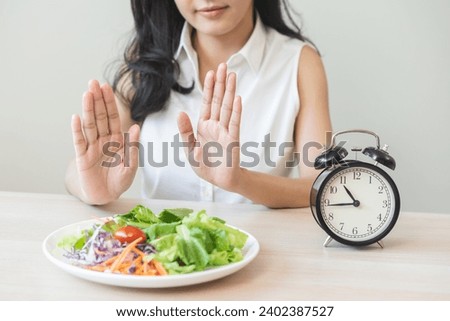 Intermittent fasting with clock, asian young woman dieting hand refuse waiting time to eat low carb food, green vegetable salad on plate. Eat food healthy first meal on brunch, lunch on table at home. [[stock_photo]] © 