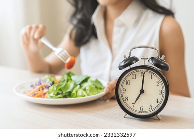 Intermittent fasting with clock, health asian young woman, girl weight loss, eating green fresh vegetable salad on dish, plate with eat healthy of breakfast food in morning, lunch on a table at home.