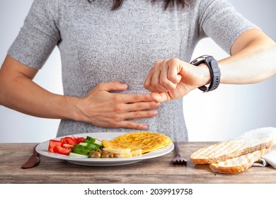 Intermittent fasting concept with a woman sitting hungry in front of food and looking at her watch to make sure she breaks fast on the correct time. A dietary modification for healthy lifestyle.