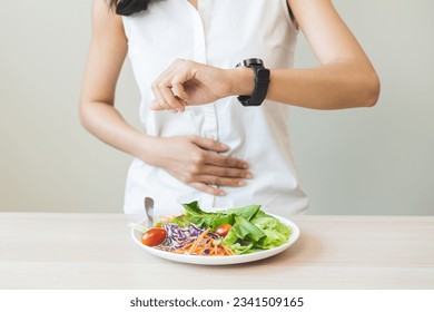 Intermittent fasting, health care asian young woman dietary, having stomach ache, temptation hungry of breakfast food in morning on table at home, looking at watch on her wrist but not yet time to eat - Shutterstock ID 2341509165