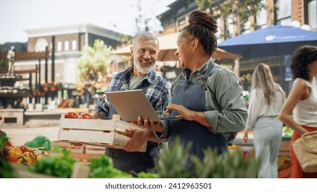 International Adult Partners Working at a Farmers Market. Diverse Couple Selling Ecological Fruits and Vegetables From an Outdoors Food Stand. Adult Female Using Laptop to Communicate with Suppliers - Powered by Shutterstock