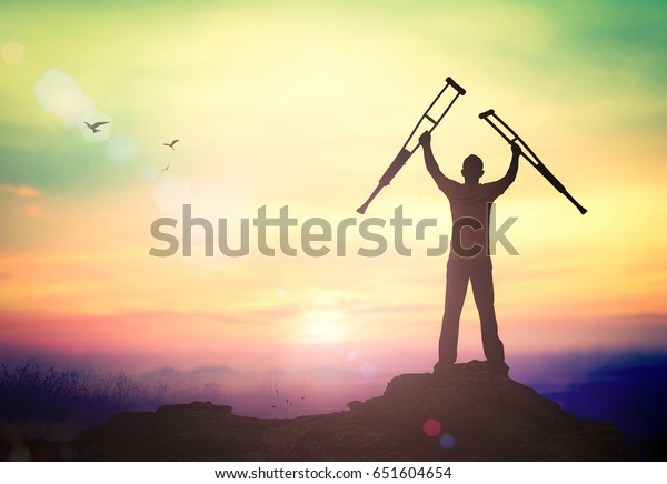 International Day of Persons\
with Disabilities (IDPD) concept: Silhouette a disabled man\
standing up and raising his crutches over mountain autumn sunset\
background