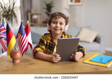 International Education For Kids. Close Up Portrait Of Adorable Little Boy Wearing Headset Enjoying Online Lesson On Digital Tablet, Sitting At Desk With Various Flags, Smiling To Camera, Free Space