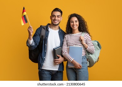 International Education. Middle Eastern Student Couple With Backpacks Holding German Flag, Portrait Of Smiling Arab Man And Woman With College Workbooks Posing On Yellow Studio Background, Copy Space