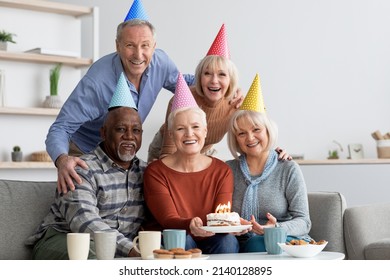 International group of elderly people happy men and women wearing colorful paper hats having birthday party at home, smiling caucasian senior lady holding birthday cake with lit candles, copy space
