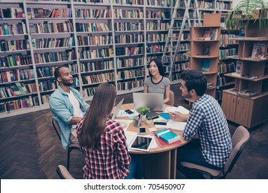 International Group Of Four Focused Clever Young Students Bookworms Studying In The Ancient University Library, Sit At The Table With Books And Devices, Talk, Discuss The Project, In Casual Outfits