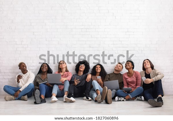 International group of friends with\
smartphones, laptops and digital tablets looking up on copy space,\
sitting on floor over white wall. Cheerful multiracial students\
using gadgets, studio\
background