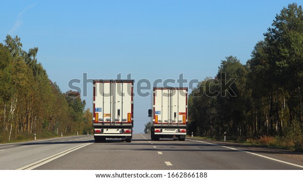 International logistics,\
shipping goods, road transportation - white long vehicle van semi\
trucks overtaking moving on a two-lane asphalted country road at\
autumn day, rear\
view