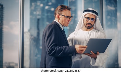 International Operations Manager Meeting Saudi Business Partner in Traditional Kandura. They're Standing in Modern Office, Using Laptop Computer. Successful Saudi, Emirati, Arab Businessman Concept.