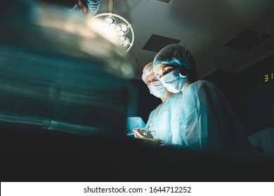 An International Professional Team Of Surgeon, Assistants And Anesthesiologist Perform A Complex Operation On A Patient Under General Anesthesia. Dark Atmospheric Photography Theme In Low Key.