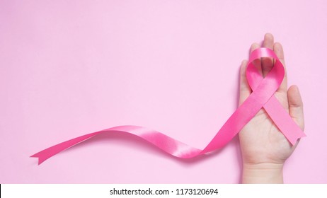 International symbol of Breast Cancer Awareness Month in October. Close up of female hand holding satin pink ribbon awareness on pink background w/ copy space. Women's health care and medical concept.
