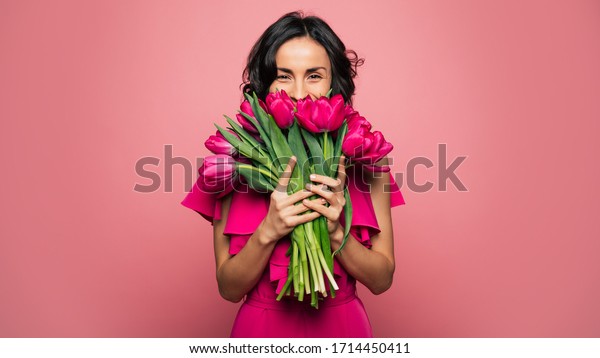 International Women\'s Day. Extremely happy woman\
in a bright pink dress is smelling a bunch of spring flowers, which\
she is holding in her\
hands.