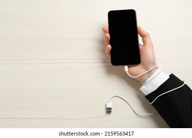 Internet addiction. Top view of man holding phone at wooden table, hand tied to device with charging cable