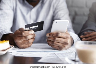Internet banking, e-commerce and online trading concept. Cropped view of African-American businesman holding mobile phone and credit card while paying bill at cafe. Selective focus on man's hands
