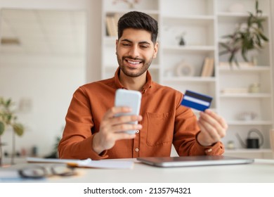 Internet Mobile Shopping Concept. Positive Middle Eastern Man Using Cell Phone Looking At Screen Holding Credit Card Purchasing Things Online Sitting At Desk At Home Office, Buying Gifts In Web Store