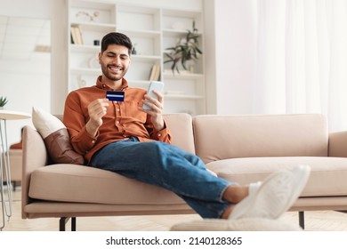 Internet Mobile Shopping Concept. Positive Middle Eastern Male Using Cell Phone Holding Looking At Credit Card Purchasing Things Online Sitting On Sofa Buying Gifts In Web Store, Full Body Length