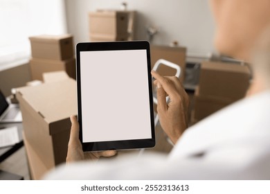 Internet shop manager using tablet computer in storage room. Empty screen copy space close up with paper containers, stacked boxes in warehouse office in background