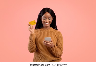 Internet Shopping Concept. Positive Black Woman With Cellphone And Credit Card Purchasing Things Online Over Pink Studio Background. Happy African American Lady Buying Gifts On Web