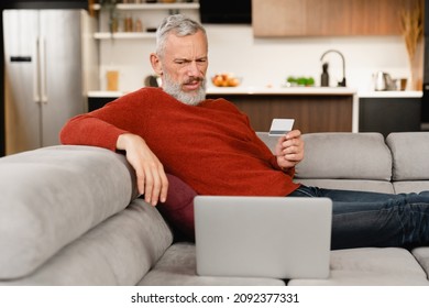 Internet theft blackmailing fraud online. Doubting caucasian mature man paying with credit card online, doing shopping , e-banking, e-commerce at home