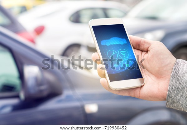 Internet of things (IOT) mobile app in smart\
phone for modern car. Hand holding smartphone controlling ADAS\
system. Vehicles parked in the\
background.