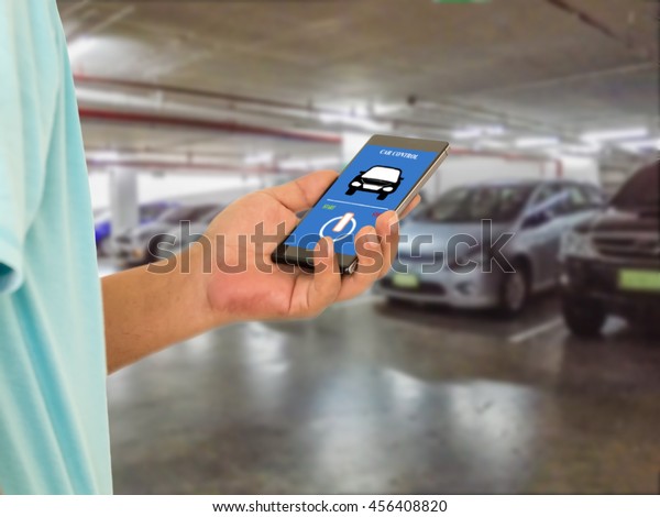 Internet of things and smart car concept. Hand\
holding smart phone with car control application on screen against\
blur car park\
background