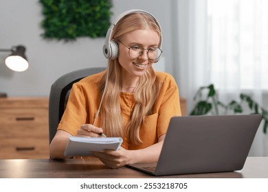 Interpreter in headphones taking notes while having video chat via laptop at table indoors