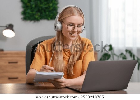 Interpreter in headphones taking notes while having video chat via laptop at table indoors
