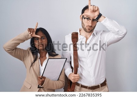 Interracial business couple wearing glasses making fun of people with fingers on forehead doing loser gesture mocking and insulting. 