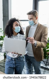 Interracial Business People In Medical Masks Talking While Standing With Laptop In Office