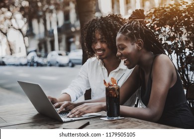 Interracial couple: black laughing girl and Asian smiling guy are looking through funny photos using the laptop while sitting in a street cafe, the glass of ice tea and the smartphone near the girl - Powered by Shutterstock