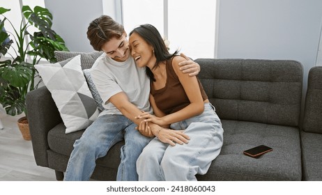 Interracial couple cuddling joyfully on a gray sofa inside a cozy living room, evoking love and togetherness. - Powered by Shutterstock
