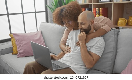 Interracial couple cuddling on a sofa while looking at a laptop in a cozy living room - Powered by Shutterstock