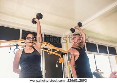 An interracial couple does a set of standing one arm overhead dumbbell tricep extensions. A husband and wife working out at the gym and keeping fit.