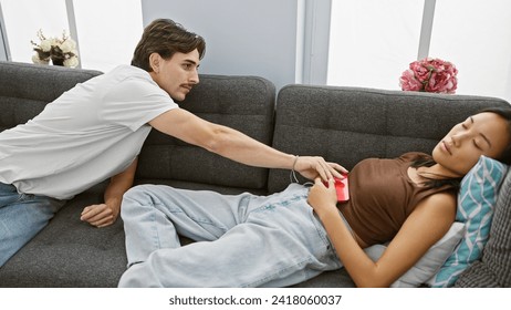 Interracial couple at home, man reaching for woman's phone while she sleeps on the couch, depicting trust and privacy. - Powered by Shutterstock