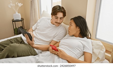 Interracial couple lounging in a bedroom with a man holding a tablet and woman using a smartphone, representing relaxation and connection. - Powered by Shutterstock