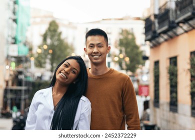 Interracial couple portrait: Asian man and Latin American woman embracing looking at camera smiling. They are standing on the city street wearing casual clothes. - Powered by Shutterstock