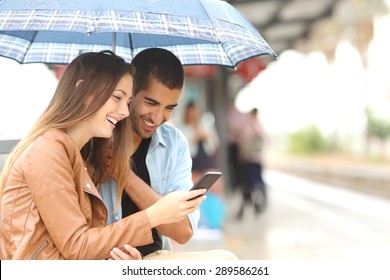 Interracial couple sharing a phone in a train station while wait under an umbrella in a rainy day - Powered by Shutterstock