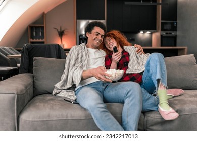 Interracial couple sitting on couch, snuggled in blankets, eating popcorn and scrolling through movies on television while talking - Shutterstock ID 2342217013