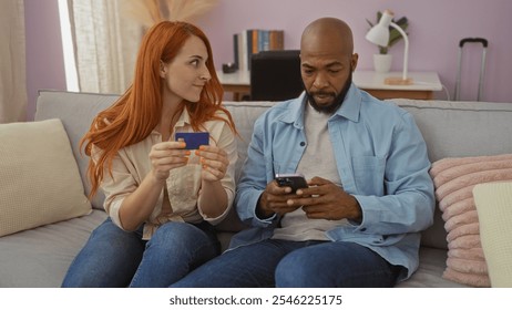 Interracial couple sitting on a sofa in a cozy living room with a woman holding a credit card and a man using his smartphone, depicting a modern relationship and shared moments at home. - Powered by Shutterstock