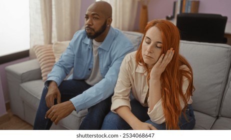 Interracial couple sitting together indoors in a living room, woman with hand on face, both looking thoughtful - Powered by Shutterstock