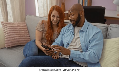 Interracial couple sitting together on a living room couch sharing a moment of joy while looking at a smartphone indoors - Powered by Shutterstock