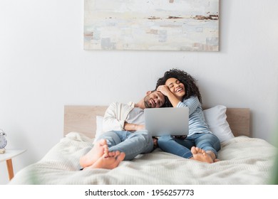 Interracial Couple Smiling And Chilling On Bed While Watching Movie On Laptop