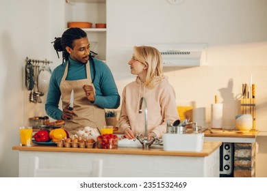 An interracial couple is standing in a kitchen and chatting while cooking lunch and washing dishes in a sink. An Arabic husband is making a veggie lunch while his wife is washing dishes. - Powered by Shutterstock