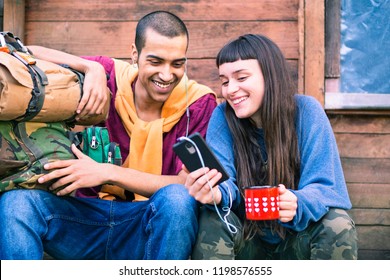 Interracial couple of young hikers looking at phone holding coffee - Happy teens on trekking outfit using smartphone sitting outdoor - Pretty girl showing video at friend - Mobile technology concept