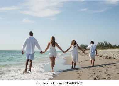 Interracial Family Of People On Vacation Walking Away Together Holding Hands On A Beach On A Sunny Day In Florida In The Water At Water's Edge With Blue Sky